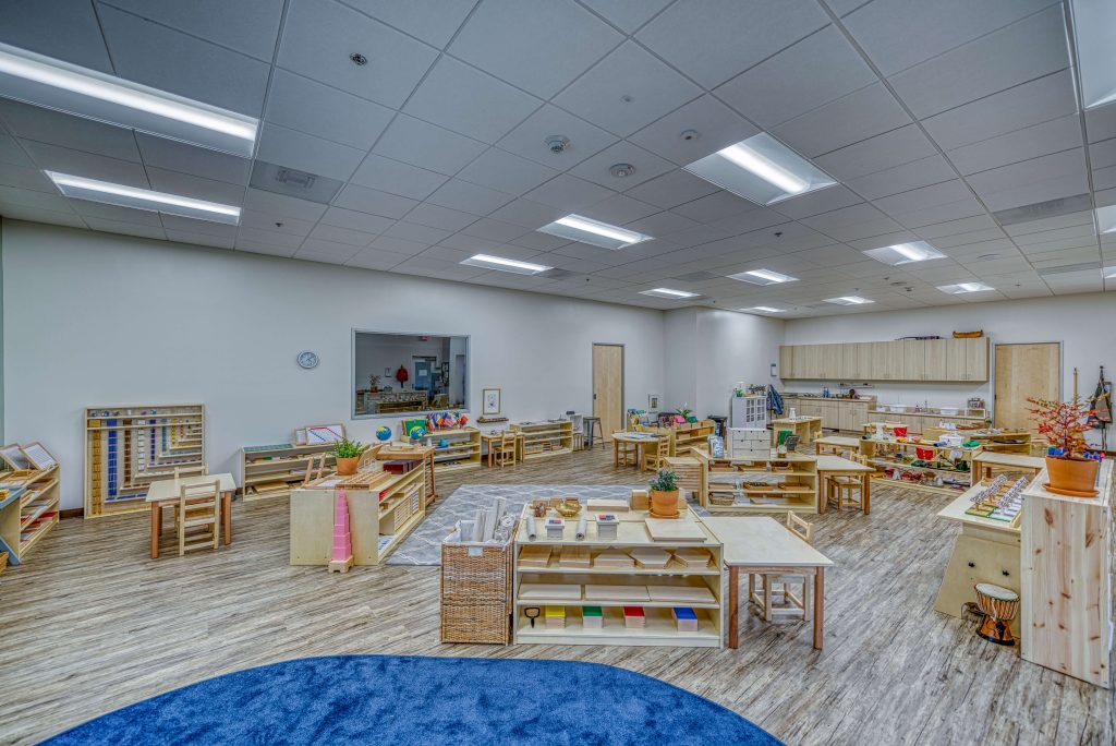 Spacious Montessori primary classroom with wooden shelves, colorful learning materials, and a large blue rug at Guidepost Montessori.