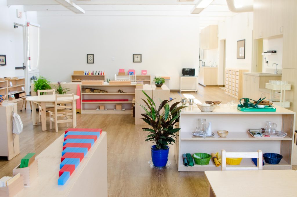 A bright Montessori Children’s House classroom featuring low wooden shelves with Practical Life and Sensorial materials, including pouring trays, glass pitchers, geometric solids, cylinders, and red and blue number rods. Plants, natural light, and child-sized furniture create an inviting, orderly environment.