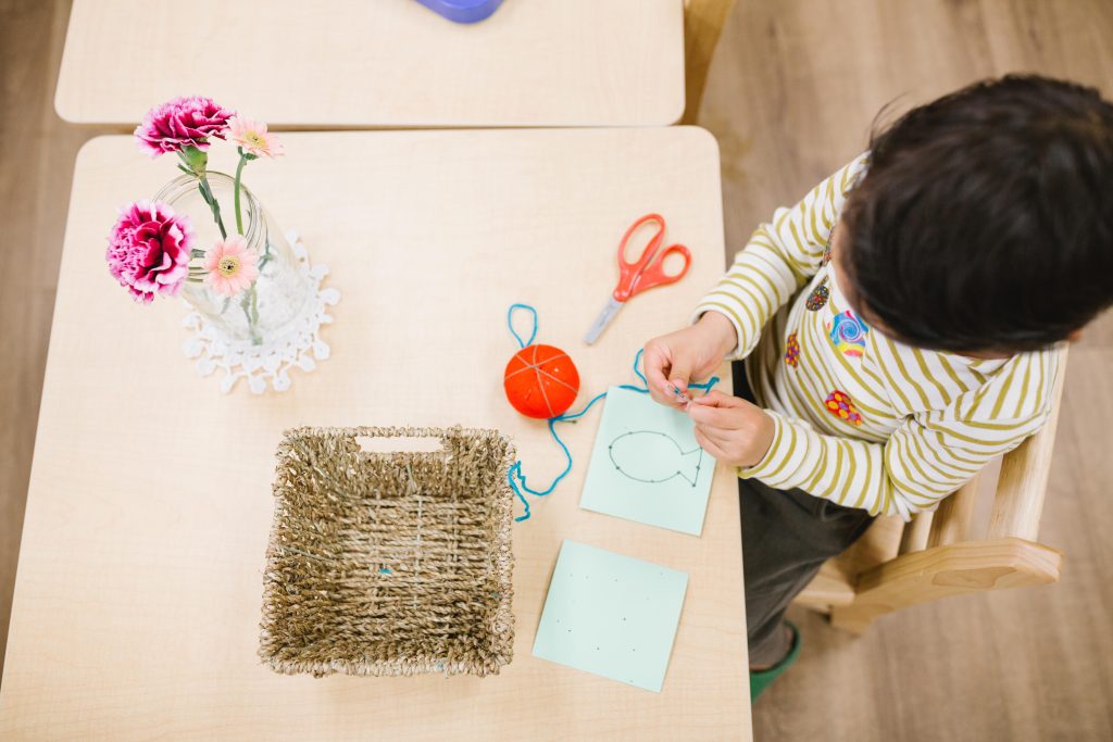 A child at a Montessori table threads yarn through a lacing card with scissors nearby, practicing coordination and focus.