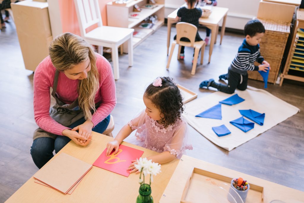 Child tracing a sandpaper letter at a small table with a Montessori guide observing and supporting.