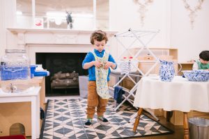 Young child in a Montessori classroom carefully folding a cloth as part of a practical life activity. Drying rack and water materials are visible in the background.