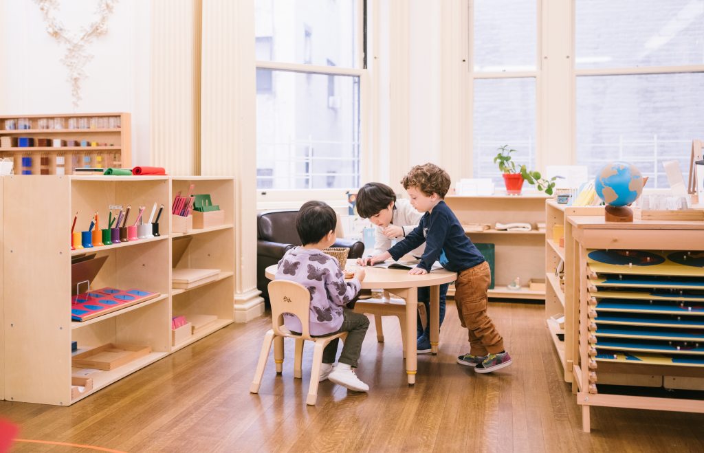 Group of young children working together around a small table in a Montessori classroom with shelves of materials in the background.