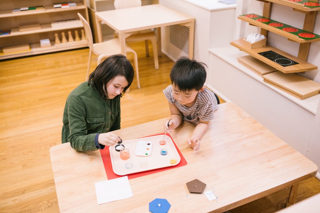 Two children working together at a Montessori activity tray, using droppers to transfer colored water during a practical life exercise.