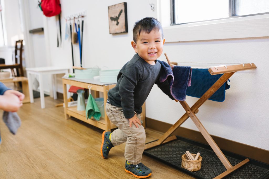 Toddler smiling while placing a cloth on a drying rack during a Montessori practical life activity.