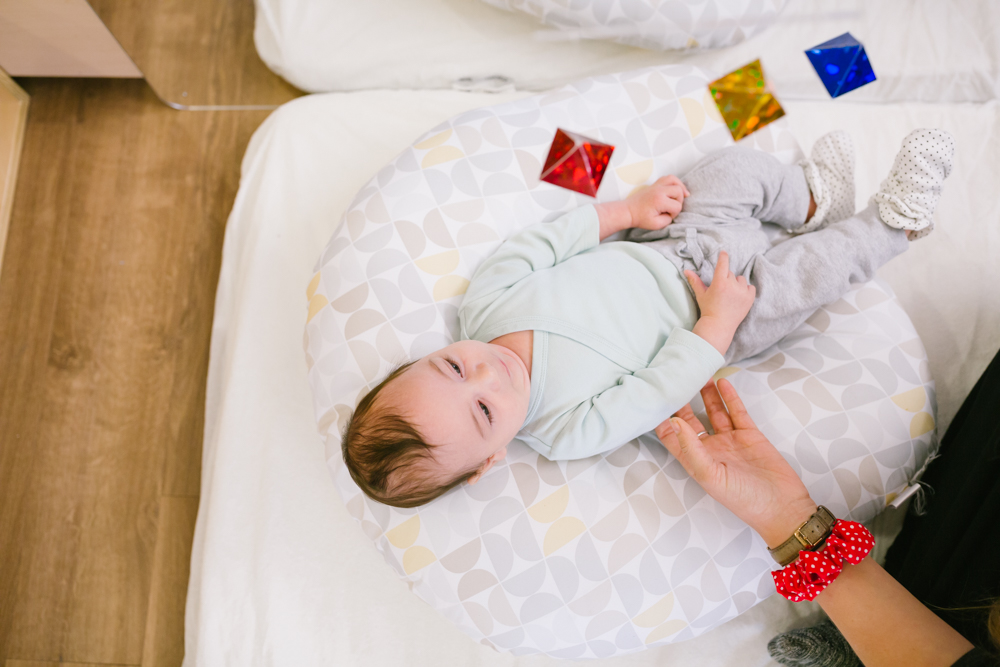 Infant lying on a floor mattress under a wooden Montessori play gym with hanging mobile toys.