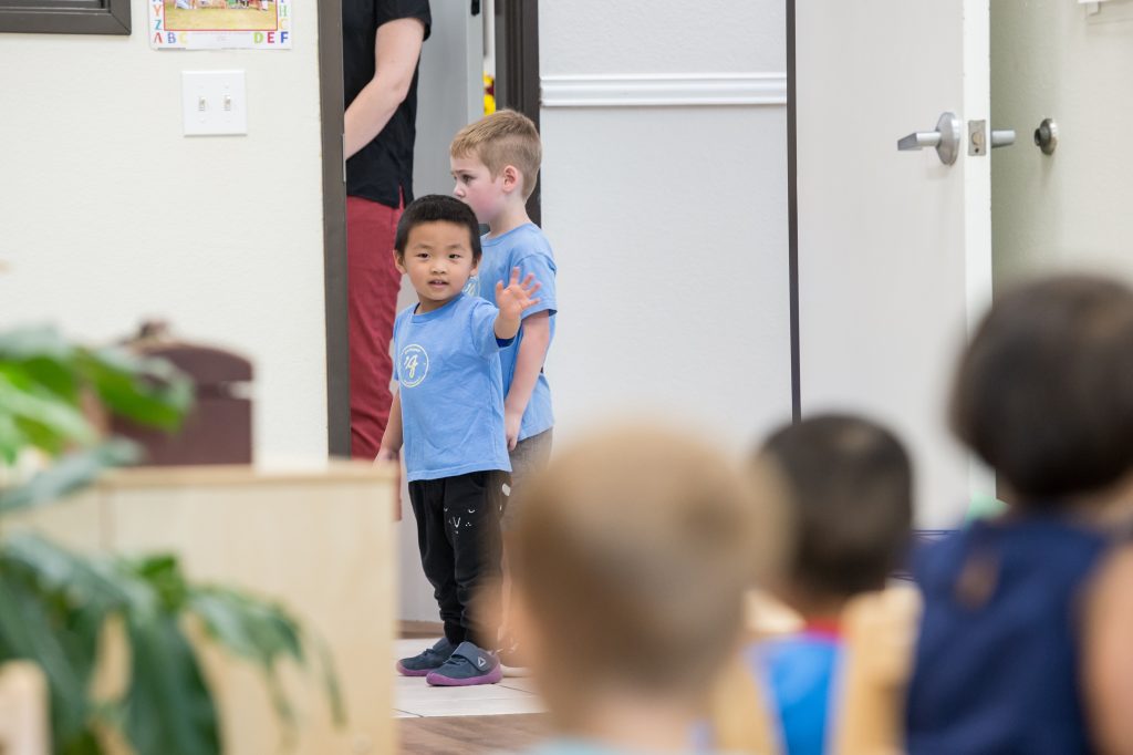 Young children enter a Montessori classroom during the holiday season, with one child pausing to wave as the day begins.