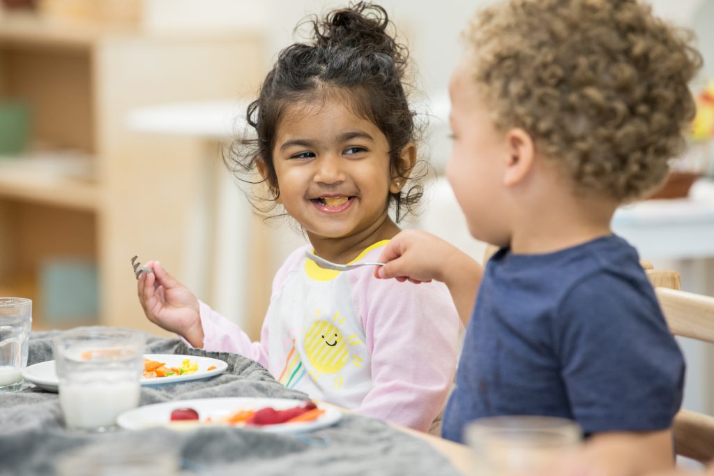 Two young children smile and connect while eating together in a Montessori classroom during the holidays.