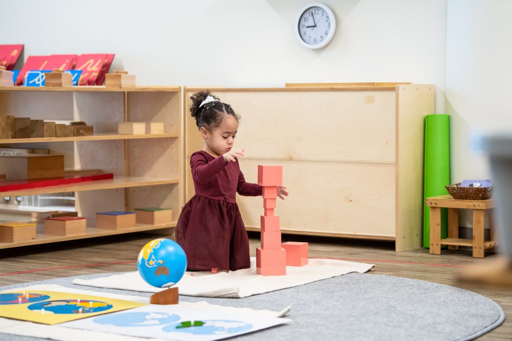 Child building a tall structure with Montessori Pink Tower cubes in a classroom.