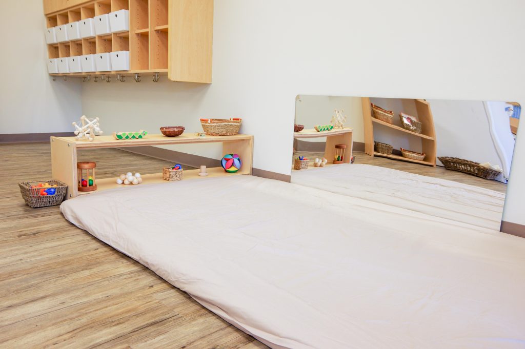 Montessori Nido classroom with a floor bed, low shelves of infant materials, and a wall mirror supporting early movement and independence.