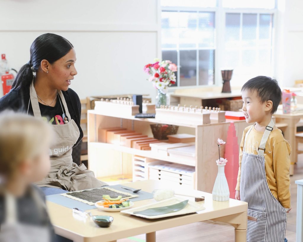 Guide at Guidepost Montessori Mahwah speaks with a smiling preschooler in an apron beside a sushi-making setup during a cultural practical life lesson.
