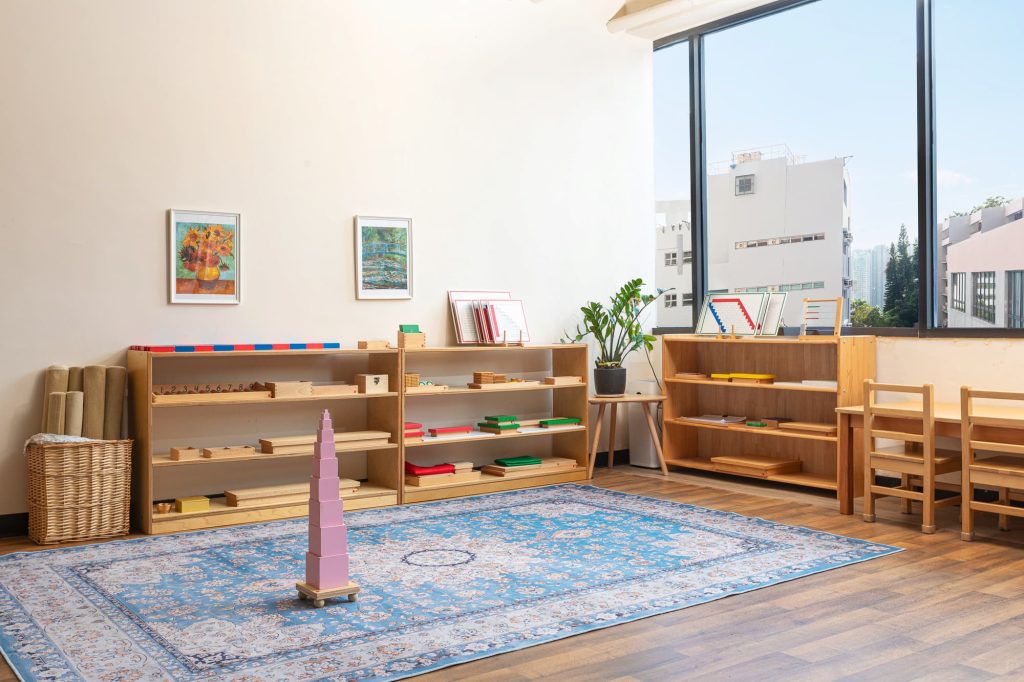 Montessori classroom with neatly arranged learning materials on low wooden shelves, a blue patterned rug, and large windows overlooking city buildings.