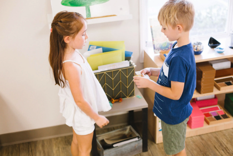 Two Montessori children standing face to face in a classroom, practicing a social interaction lesson with one child offering an item to the other.