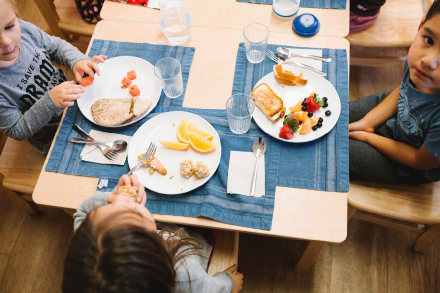 Children sitting together at a small table in a Montessori classroom, eating snacks on placemats with real plates, cups, and utensils.