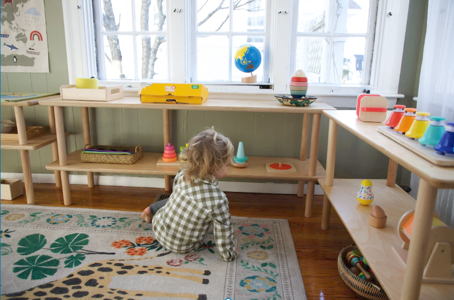 Toddler sitting on a rug independently choosing Montessori materials from low wooden shelves in a calm, light-filled classroom.