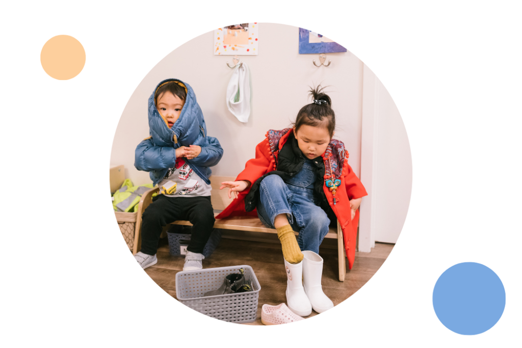Two young children independently putting on their coats and shoes in a Montessori classroom entry area. One child is zipping a blue jacket while the other is pulling on white boots, practicing self-care and independence.